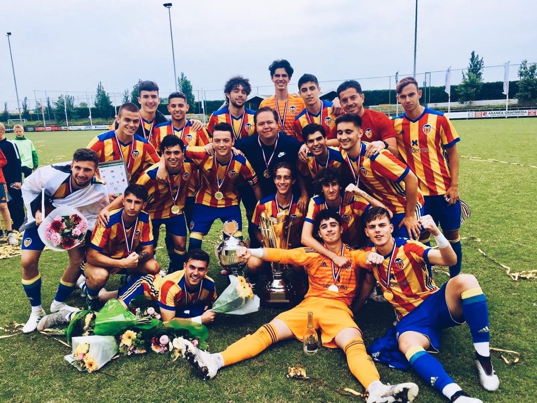 Valencia CF youth players in red and yellow striped jerseys celebrating with medals, flowers and a trophy, with Jaime González Albarrán in the middle of the team.